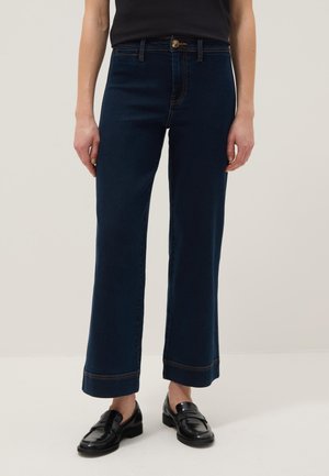 Persoon die donkerblauwe wijde spijkerbroek, zwarte loafers en een zwart topje draagt, staand tegen een effen lichte achtergrond.