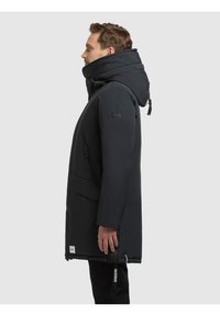 Parka neagră izolată, cu guler înalt și glugă ajustabilă, având un buzunar lateral cu fermoar și detalii de marcă. Textură netedă, mată.