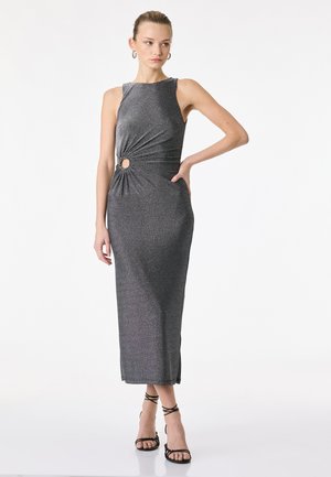 Frau trägt ein ärmelloses Midi-Kleid in metallischem Grau mit seitlichem Ausschnitt und runder Details, kombiniert mit schwarzen, römisch inspirierten High-Heels-Sandalen.