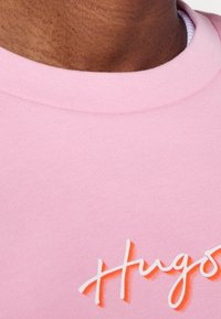 Roze katoenen sweatshirt met een ronde hals en een verhoogd wit tekstlogo "Hugos" rechtsonder. Zachte textuur en casual ontwerp.