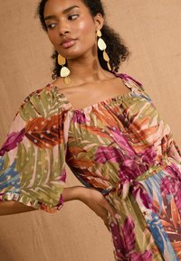 Florale maxi-jurk met pofmouwen, in levendige kleuren zoals roze, groen en blauw, versierd met een strikceintuur en gouden accenten.