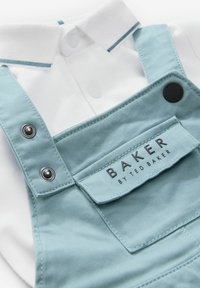 Svijetloplave kombinezone sa srebrnim metalnim dijelovima, ispod kojih se nalazi teksturirana bijela majica. Vidljiva oznaka brenda "BAKER by TED BAKER".