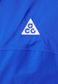 Modra tkanina z gladko teksturo ima bel trikotni logotip s "acg" znotraj. Šivanje se vidi ob spodnjem robu.