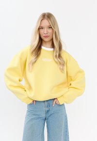 Żółta, przew oversize'owy sweatshirt z białym kołnierzykiem i logo "MIRONS". Materiał jest miękki, z szerokimi rękawami i luźnym krojem, w połączeniu z jasnymi dżinsami.
