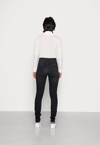 Jeans skinny de mezclilla negra con cintura alta, cinco bolsillos y costuras en contraste, combinados con una camiseta blanca de manga larga y zapatos.
