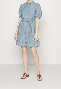 Světle modré džínové šaty s krátkými nabíranými rukávy, předním zapínáním na knoflíky a zavázaným páskem v pase. Mají tiered volánkový lem a měkkou texturu.