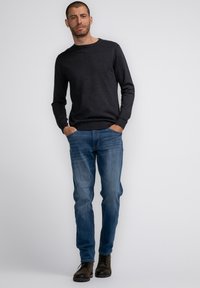 Dark grey sweater with a round neckline, blue denim jeans, and brown lace-up boots. Model stands with hands in pockets against a light background.