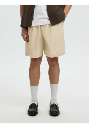 Uomo che indossa pantaloncini beige, calzini bianchi, mocassini neri, maglietta bianca e camicia marrone a maniche corte con le mani nelle tasche.