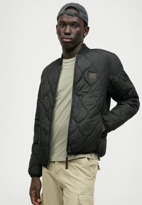 Schwarze gesteppte Bomberjacke mit Reißverschluss, gerippten Bündchen und einem braunen Lederlogo-Patch; kombiniert mit einem hellgrünen Hemd.