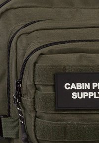 Mochila verde oliva hecha de tela duradera, con compartimentos con cremallera, una etiqueta de parche y acentos de panel texturizado.