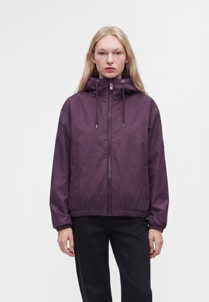Junge Frau mit langen blonden Haaren, die eine lila Kapuzenjacke mit Reißverschluss und schwarze Hose trägt, steht vor einem einfarbigen weißen Hintergrund.