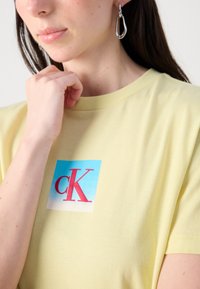 Sárga pamut póló, kék és piros grafikával, amelyen a „cK” betűk találhatók a mellkason. A modellen ezüst csepp fülbevaló van.