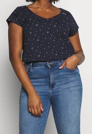 Vrouw draagt een marineblauwe korte mouwen top met kleine witte hartjes, ankers en sterren, gecombineerd met blauwe jeans met een normale taillehoogte.