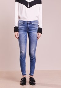 Vaqueros ajustados azules con ligero desgaste, combinados con una blusa en blanco y negro que presenta un cuello con cremallera y mangas en contraste. Zapatillas negras sin cordones.