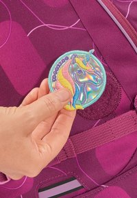 Sac à dos violet avec une texture de tissu lisse et un patch holographique de licorne circulaire en cours d'application. Les accents comprennent des détails roses et bleus.