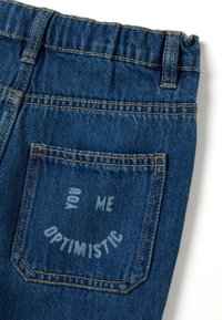 Vaqueros de denim de color azul, con un bolsillo trasero que lleva impreso el texto "YOU ME OPTIMISTIC" y detalles de costura amarilla.