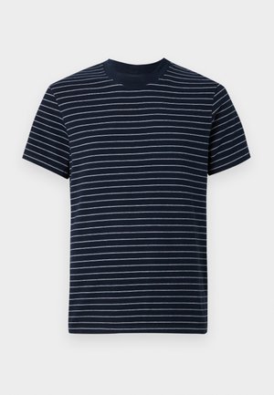Ung mand med krøllet hår iført en sort og hvid stribet t-shirt, mørke jeans og sorte sko, stående op ad en ensartet lys væg.