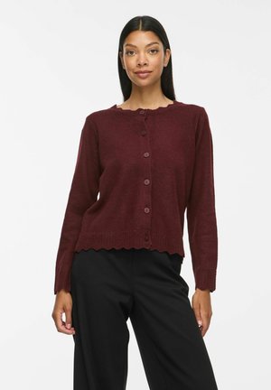 Femme portant un cardigan bordeaux à manches longues, boutonné avec des bords festonnés et un pantalon noir sur fond blanc.
