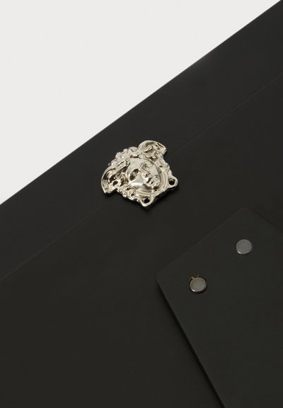 Emblème tête de Méduse en argent fixé à une surface noire avec une plaque noire rectangulaire et deux rivets métalliques à proximité.