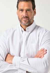 Weißes Hemd mit Knopfleiste aus strukturiertem Stoff, mit spitzem Kragen und einer Brusttasche, die Arme verschränkt, neutraler Hintergrund.