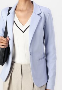 Ljusblå blazer med en strukturerad design, enkel knappstängning och bröstficka, matchad med en krämfärgad tröja och beige byxor.