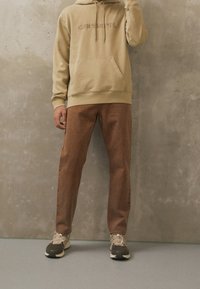 Beige huppari, jossa on brodeerattu logo, etutasku, yhdistettynä ruskeisiin suoriin housuihin ja vaaleanruskeisiin lenkkikenkiin, taustalla betoniseinä.