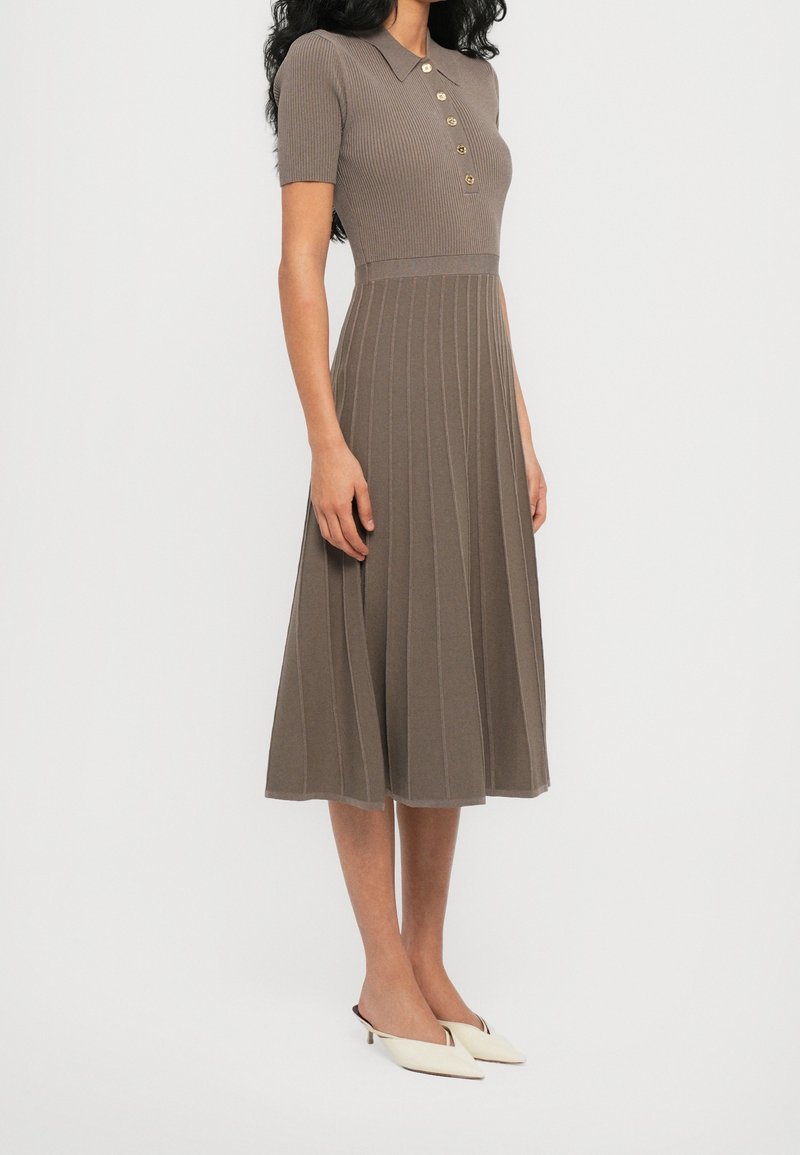 Femme portant une robe midi en maille côtelée marron, manches courtes, col boutonné, et des chaussures blanches à talons bas pointus.