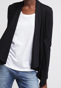 Svart, lätt öppen cardigan med långa ärmar över en vit t-shirt med rund halsringning. Matchad med blå jeans. Mjuk tygstruktur.