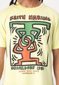 Yellow cotton T-shirt featuring bold black outlines of stylized figures in orange and green, with text reading "KEITH HARING DÜSSELDORF 1981."