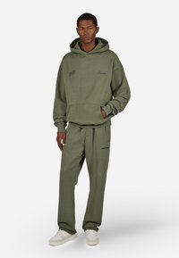 Oliwkowa bluza z kapturem z kieszenią kangurkową i haftowanym logo, w zestawie z dresowymi spodniami w tym samym kolorze, które mają boczne kieszenie i czarne akcenty tekstowe.