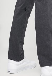 Grijze broek met een subtiele textuur, voorzien van een zwart logo aan de zijkant, gecombineerd met witte sneakers met een opvallend ontwerp.