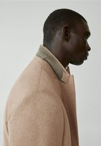 Beige wollen jas met een gestructureerde oppervlakte, voorzien van een groene kraag. Klassiek ontwerp met een getailleerde pasvorm en strakke lijnen.