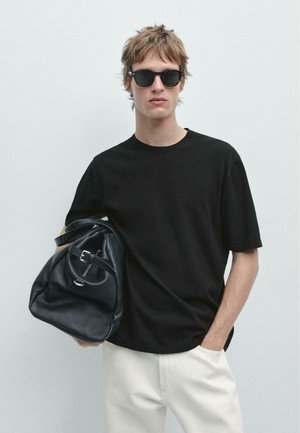 Junger Mann mit schwarzen Sonnenbrillen und einem schwarzen T-Shirt, hält eine schwarze Ledertasche mit einer Hand, steht vor einem schlichten hellen Hintergrund.