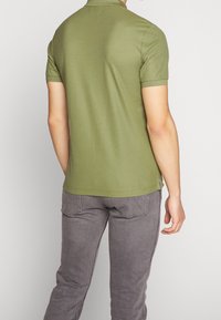 Polo vert olive à manches courtes, fabriqué dans un tissu lisse. Présente un col discret et un ourlet droit. Porté avec un pantalon gris.