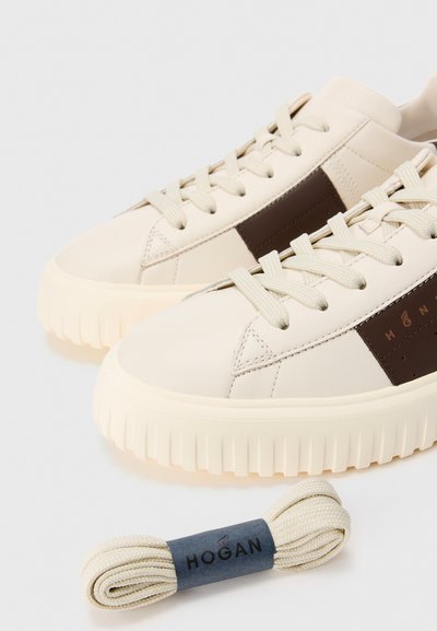 Un paio di sneaker beige con striscia laterale marrone e dettagli del brand, lacci attaccati e un set extra di lacci beige con un braccialetto del marchio.