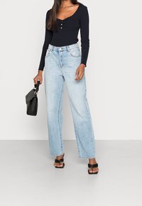 Jean denim long bleu clair à taille haute, associé à un haut côtelé bleu marine avec un décolleté festonné et des boutons. Tenant un sac à main noir.