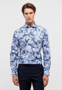 Camicia da uomo a maniche lunghe con motivo paisley blu e bianco, realizzata in tessuto liscio con colletto a punta e chiusura a bottoni. Pantaloni scuri indossati sotto.
