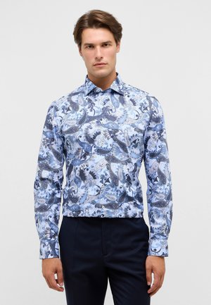 Blauw-witte paisley patroon overhemden met lange mouwen, gemaakt van een gladde stof met een puntige kraag en knoopsluiting. Donkere broek eronder gedragen.