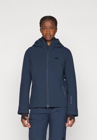Chaqueta impermeable de color azul marino con capucha, cierre de cremallera frontal y bolsillos laterales. Presenta un logo en el pecho y puños elásticos con orificios para los pulgares.