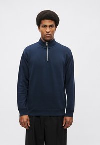 Tmavomodrá mikina na zips s dlhými rukávmi, rebrovanými manžetami a rovný lemom. Vyrobená z hladkého materiálu, s strieborným zipsom.