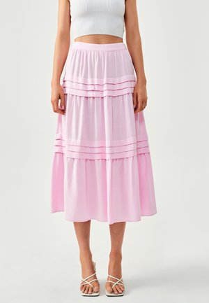 Mujer con falda midi rosa claro de volantes, top corto blanco acanalado y sandalias blancas de tacón con tiras, posando sobre un fondo liso.