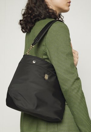 Femme portant une veste à motif pied-de-poule vert, portant un grand sac noir avec une sangle en cuir et un emblème doré sur l'épaule, sur un fond uni.
