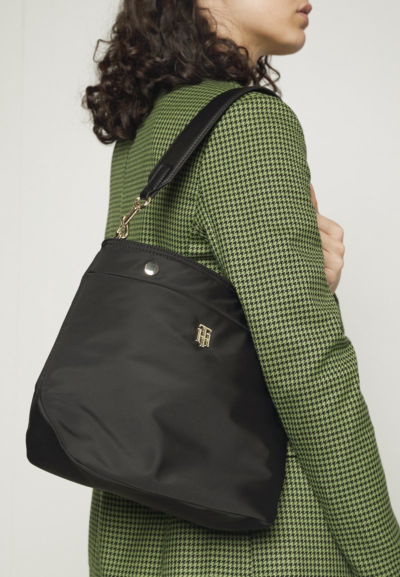Femme portant une veste à motif pied-de-poule vert, portant un grand sac noir avec une sangle en cuir et un emblème doré sur l'épaule, sur un fond uni.