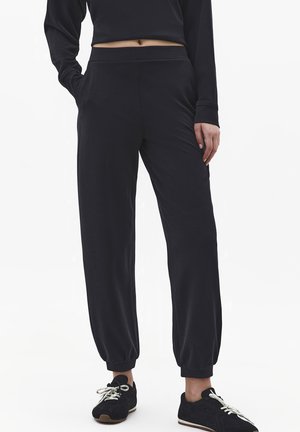 Czarne spodnie dresowe z elastycznymi ściągaczami noszone z czarnymi trampkami, połączone z czarnym krótkim topem z długim rękawem, modelka stoi z jedną ręką w kieszeni.