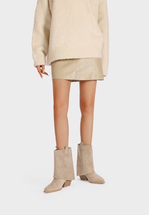 Gambe con minigonna in pelle beige, maglione oversize crema morbido e stivali beige in camoscio a metà polpaccio con tacchi in legno su sfondo bianco.