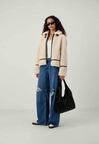 Bežinė kailinė striukė su juodu apvadu, laisvi mėlyni suplyšę džinsai ir juodas plisuotas rankinukas. Modelis dėvi juodus saulės akinius.