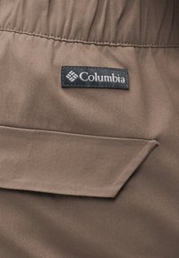 Καφέ ύφασμα με καπάκι τσέπης και μια μαύρη υφαντή ετικέτα ραμμένη από πάνω, που εμφανίζει το λογότυπο της μάρκας "Columbia" με λευκή γραμματοσειρά.