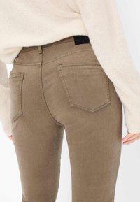 Bruine slim fit jeans met achterzakken. De stof heeft een zachte textuur en een lichte glans, gecombineerd met een lichtgekleurde trui.