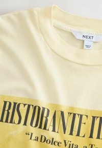 Geel katoenen t-shirt met korte mouwen. Voorzien van een grote zwarte bedrukte tekst "RISTORANTE IT" op de voorkant en een ronde halslijn.