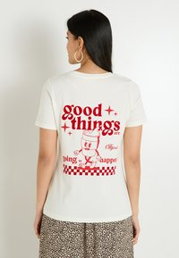 T-shirt couleur crème avec un texte graphique rouge et un personnage de dessin animé au dos, accompagné d'un motif à carreaux en bas.